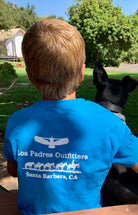 Back view of boy sitting outside wearing the comfortable blue cotton Kids Short Sleeve Tee with the Los Padres Outiftters logo on the back