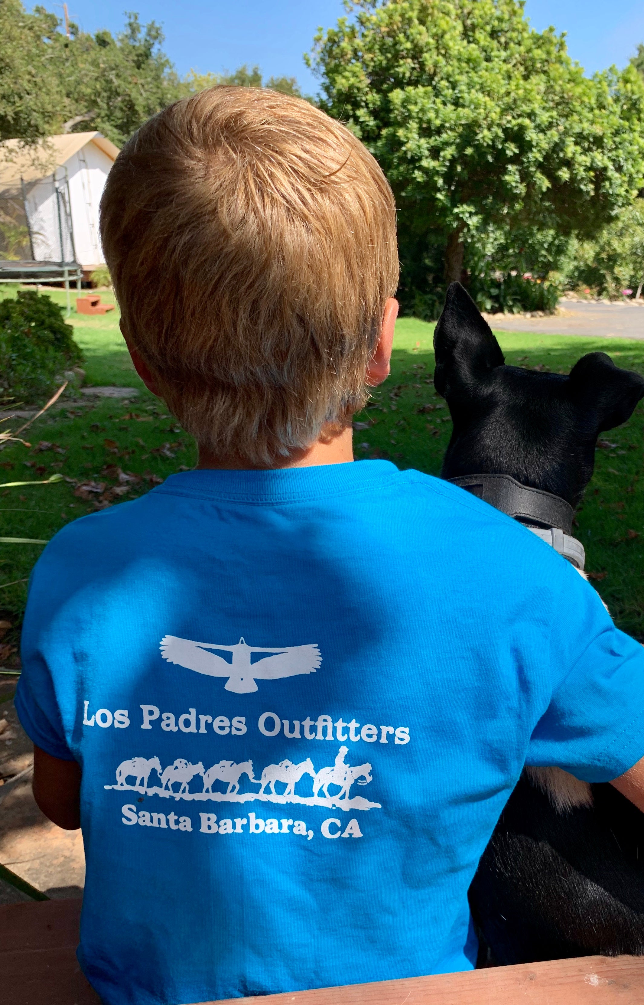 Back view of boy sitting outside wearing the comfortable blue cotton Kids Short Sleeve Tee with the Los Padres Outiftters logo on the back